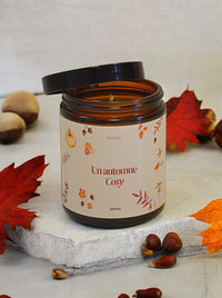 Bougie parfumée "Un Automne Cosy" éteinte,  posée sur une table basse à côté d'un plaid en laine et d'une tasse de thé fumante.  Le pot ambré et l'étiquette "Un Automne Cosy"  invitent à la détente et au cocooning