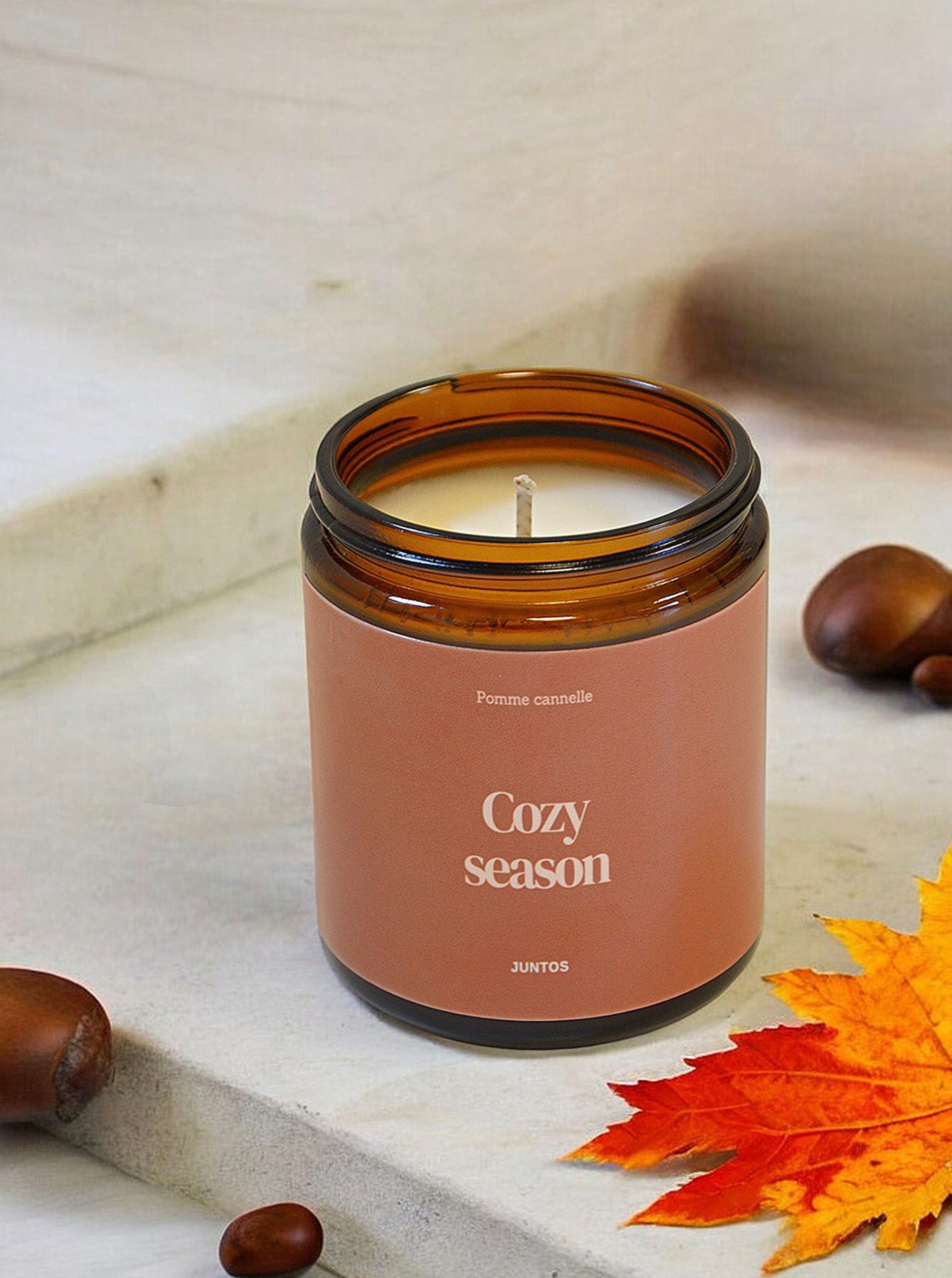 Gros plan sur la bougie parfumée "Cozy Season" entourée d'éléments cocooning :  un plaid en laine, des chaussettes moelleuses et une tasse de chocolat chaud. Le pot ambré et l'étiquette waterproof mettent en valeur le parfum [Nom du parfum] et l'ambiance chaleureuse.