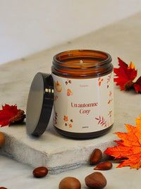 Bougie parfumée "Un Automne Cosy" allumée,  diffusant une lumière douce et chaleureuse dans un salon décoré avec des feuilles d'automne.  Le pot ambré scintille et l'étiquette "Un Automne Cosy"  ajoute une touche élégante à l'ambiance.